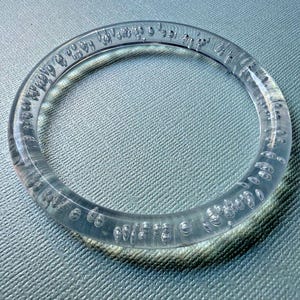 Könnte beinhalten: Ein rundes, transparentes Plastikarmband mit kleinen Luftblasen, die darin eingebettet sind. Das Armband hat eine glatte Oberfläche.