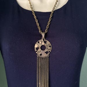 May include: A vintage gold-tone necklace with a circular pendant featuring a sunburst design. The pendant has a series of dangling chains. The necklace is displayed on a mannequin wearing a navy blue top.