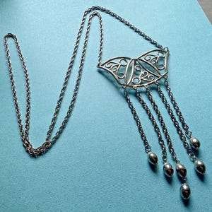 Puede incluir: Collar plateado con un colgante de filigrana decorativo y detalles de cadena colgantes. El collar presenta una cadena larga y un diseño simétrico, con adornos en forma de lágrima en los extremos de las cadenas. La joya se exhibe sobre una superficie azul.