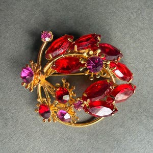 Puede incluir: Broche vintage dorado con diseño circular, adornado con piedras preciosas rojas y moradas brillantes. El broche presenta un arreglo floral, con piedras rojas ovaladas y piedras moradas redondas. Una joya.