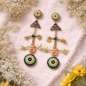 Puede incluir: Un par de pendientes hechos a mano con un diseño único. Los pendientes presentan una combinación de metal dorado, cuentas negras, rosas, verdes y amarillas. El diseño incluye elementos circulares y triangulares, con detalles colgantes.