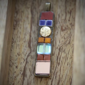 Könnte beinhalten: Ein bunter Mosaik-Anhänger mit einem goldenen Akzent. Der Anhänger besteht aus kleinen, quadratischen Fliesen in Rot-, Orange-, Blau-, Grün- und Rosatönen. Der Anhänger hängt an einer silbernen Kette.