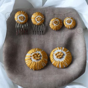 Puede incluir: Conjunto de cuatro botones de tela bordados en forma circular con un centro blanco y costuras doradas. Los botones están dispuestos en dos pares, uno en un peine plateado y el otro en ganchos de pendientes plateados.