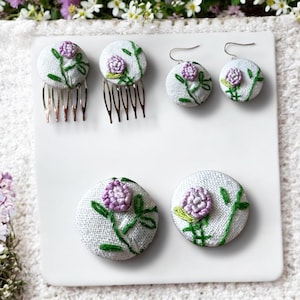 Könnte beinhalten: Set aus vier weißen Stoffknöpfen mit gestickten lila Blumen und grünen Blättern. Die Knöpfe sind an zwei silbernen Haarkämmen und zwei silbernen Ohrhaken befestigt.