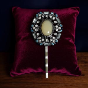 Puede incluir: Una horquilla plateada con una piedra central ovalada de color crema, rodeada de cristales transparentes y azules. La cabeza decorativa está colocada sobre un alfiler largo y fino de plata, exhibida sobre un cojín de terciopelo burdeos.