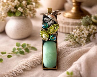 May include: A rectangular pendant with a hummingbird and leaf design. The pendant features a large, light green stone at the bottom, with green and clear crystals and a silver hummingbird at the top. The pendant is set in a gold-toned metal frame.