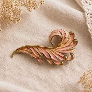 May include: A vintage-style brooch in the shape of a stylized feather. The brooch features a pink enamel finish with gold-tone metal accents. The design includes a curled tip and detailed feather-like elements.
