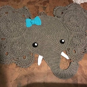 May include: A gray crocheted elephant rug with a blue bow. The elephant has a round body and large ears. The rug is made with a delicate crochet stitch.