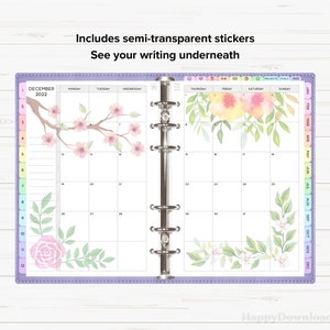 May include: A purple monthly planner with a December 2022 calendar. The planner has a clear plastic cover with a floral design. The floral design includes pink flowers, green leaves, and a pink rose.