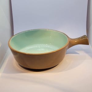 May include: A brown ceramic bowl with a light blue interior and a single handle. The bowl is designed for serving food.