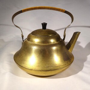 May include: A vintage brass teapot with a curved spout and a black knob on the lid. The handle is made of metal and wrapped with woven rattan. The teapot has a rounded body and a shiny, golden finish.