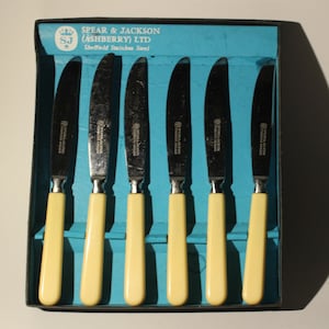 May include: A set of six stainless steel table knives with ivory-colored handles in a blue and white box. The box has the text "Spear & Jackson (Ashberry) Ltd Sheffield Stainless Steel" printed on it.