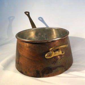 May include: A copper saucepan with a brass handle and a brass side handle. The interior is silver-colored. The pot has a rounded shape and is designed for cooking. The copper exterior has a rich, aged patina.