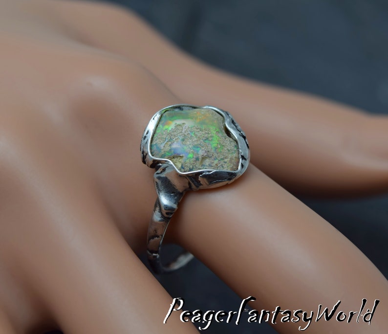 May include: A silver ring with a large, irregular shaped opal gemstone set in a textured silver band. The opal has a green, white, and brown color.
