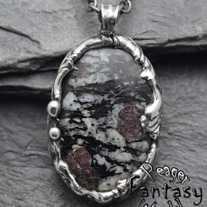 Könnte beinhalten: Ein ovaler Anhänger mit einem dunklen Stein mit weißen und schwarzen Adern sowie tiefroten Einschlüssen, gefasst in eine strukturierte Silberfassung an einer dunklen Metallkette. Der Text "Eager Fantasy World" ist unten rechts zu sehen.