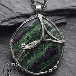 Op de afbeelding: Een zilveren ketting met een hanger met een groene en paarse edelsteen die in een zilveren frame is gezet. De edelsteen heeft een uniek, draaiend patroon.