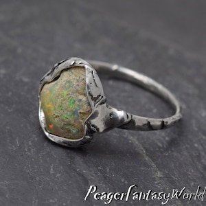 May include: A silver ring with a rough, textured band and a large, irregular-shaped opal gemstone set in the center. The opal has a milky white base with flashes of green and yellow.