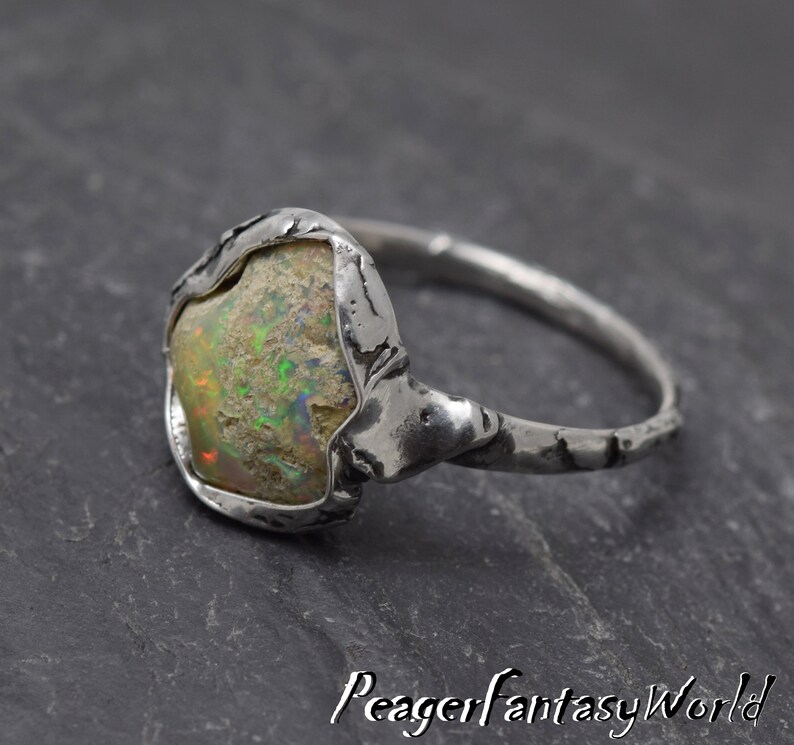 May include: A silver ring with a rough, textured band and a large, irregular-shaped opal gemstone set in the center. The opal has a milky white base with flashes of green and yellow.