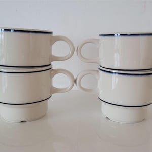Puede incluir: Cuatro tazas de cerámica blanca apiladas en pares. Cada taza tiene un borde azul oscuro alrededor de la parte superior e inferior, y un asa a juego. Las tazas están dispuestas sobre una superficie blanca.