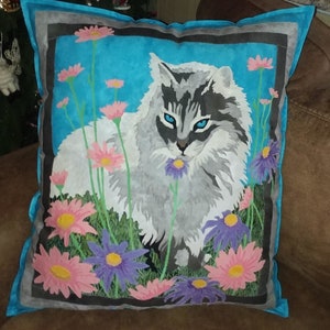 May include: A decorative pillow with a blue background and a white cat with gray markings. The cat is surrounded by pink and purple flowers.