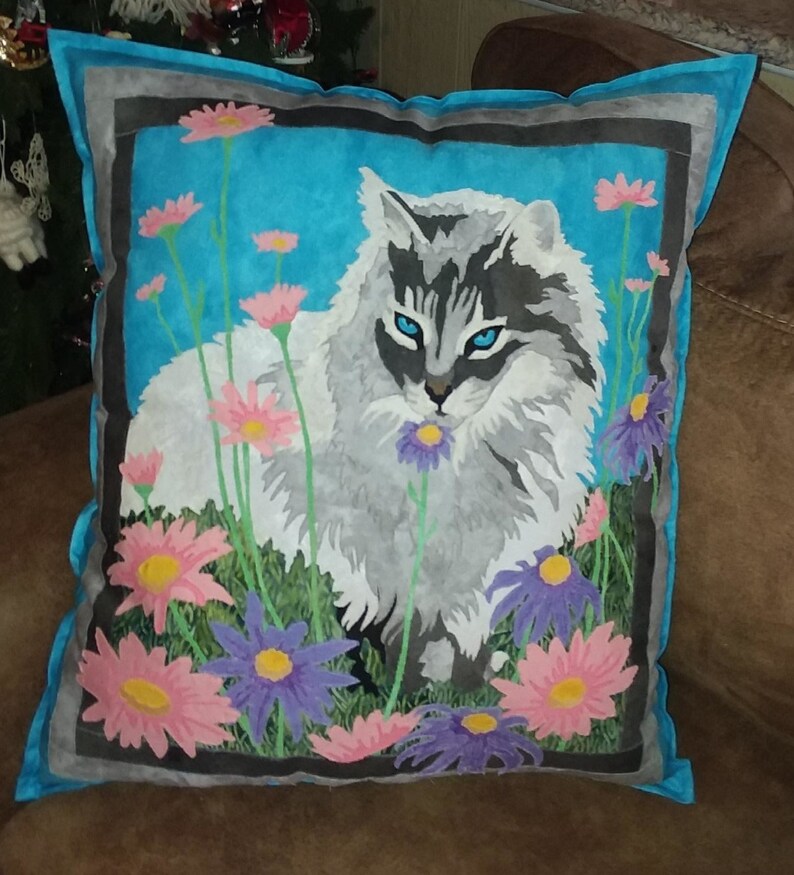 May include: A decorative pillow with a blue background and a white cat with gray markings. The cat is surrounded by pink and purple flowers.