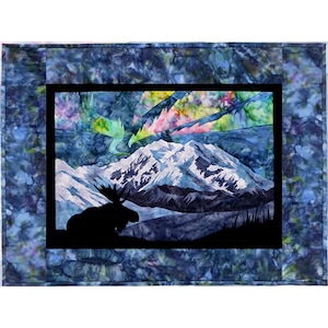 May include: A framed quilt depicting a moose silhouetted against a mountain range with a colorful aurora borealis in the sky.