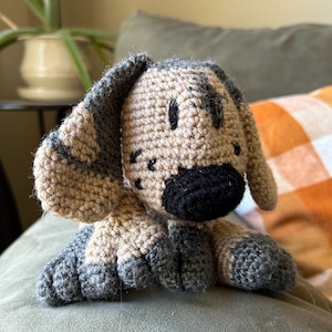 May include: A crocheted stuffed dog toy with brown and grey fur, black eyes, and a black nose. The dog is lying on a grey sofa.