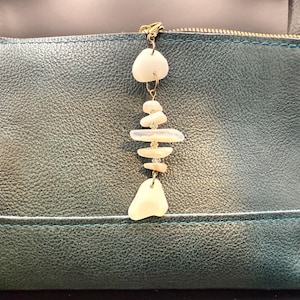 May include: A teal leather pouch with a gold zip and a decorative charm. The charm is made of white, irregularly shaped stones and small clear beads, suspended from a gold chain. The pouch has a stitched border.