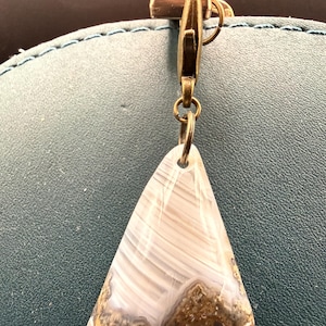 May include: A triangular pendant made of agate stone, featuring bands of white and gray. The pendant is attached to a bronze-colored clasp and ring. The stone has a brown, textured base.