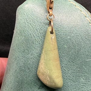 May include: A teal leather pouch with a brass zip and a stone pendant. The pendant is a light green, teardrop-shaped stone attached to the zip pull with a small brass ring. The leather has a textured appearance.