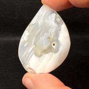 May include: A white agate stone with gray and brown banding. The stone is smooth and polished, and has a natural, organic shape.