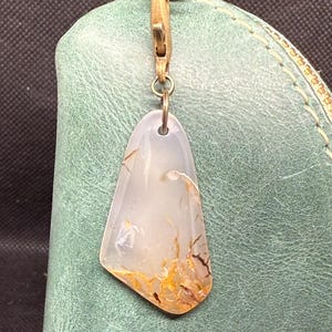 Pode incluir: Um pingente transparente em forma de gota com inclusões marrons e laranja pende de um fecho dourado. O pingente está preso a uma bolsa de couro verde-azulado com um zíper dourado.