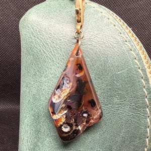 Op de afbeelding: Een bruin-amberkleurige hanger met een unieke, onregelmatige vorm hangt aan een goudkleurige sluiting. De hanger is bevestigd aan een groene leren portemonnee. De hanger heeft een gemarmerd uiterlijk met zilveren vlekken.