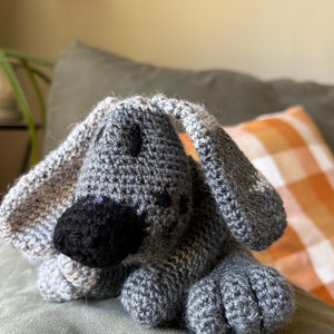 Pode incluir: Um brinquedo de cachorro de crochê cinza com olhos pretos e nariz preto. O brinquedo está deitado em um sofá cinza.