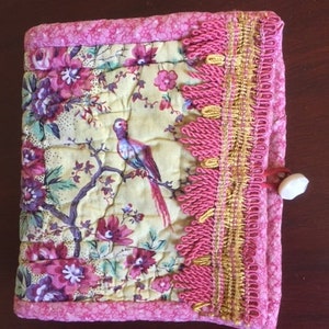 May include: A small pink fabric pouch with a floral print and a bird design. The pouch has a gold and pink trim with tassels. The pouch has a white button closure.
