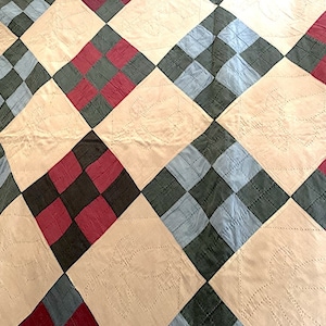 Op de afbeelding: Een quilt met een patchwork-ontwerp met vierkanten van rode, blauwe en zwarte stof. De quilt is gemaakt met een ruitpatroon en heeft een crèmekleurige achtergrond.