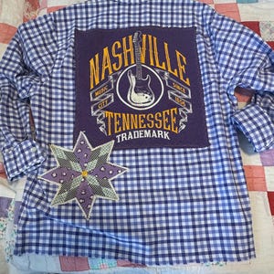 May include: Blue and white checkered shirt with a purple patch featuring "Nashville Tennessee" and a guitar graphic. A star-shaped patch with floral and geometric patterns is sewn on the lower left side. The shirt has long sleeves.