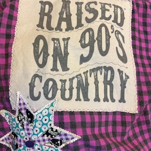 Könnte beinhalten: Ein rosa-schwarz kariertes Hemd mit einem großen weißen Aufnäher auf dem Rücken. Der Aufnäher trägt den grauen Schriftzug "RAISED ON 90'S COUNTRY". Auf der Vorderseite ist ein dekorativer Stern aufgenäht.