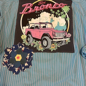 May include: A turquoise and white striped button-up shirt with a large patch featuring a pink Ford Bronco, palm trees, and the word "Bronco". A smaller, flower-shaped patch is sewn on the shirt.