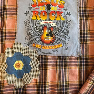 以下が含まれることがあります： ピンク、茶色、黄色のチェック柄のシャツ。 「JESUS IS MY ROCK & MY SALVATION」の文字とギターのグラフィックが描かれたパッチが付いています。 青と白の花の形をしたアップリケがシャツに縫い付けられています。