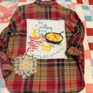 May include: A red, brown, and black plaid flannel shirt with a patch featuring a Southern-themed graphic. The graphic includes text that reads "as Southern as it gets" and "Celebrating 20 Delicious Years!" A decorative flower patch is sewn on the shirt.