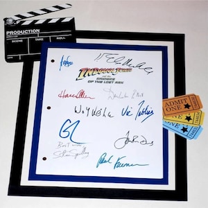 May include: A white framed print with signatures from the cast and crew of the movie "Indiana Jones and the Raiders of the Lost Ark". The signatures include Harrison Ford, Steven Spielberg, and others. The print is surrounded by a black and blue mat.