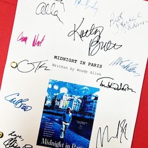 May include: A white script with multiple signatures in black, blue, and pink ink. The title "MIDNIGHT IN PARIS" is visible, along with the text "Written by Woody Allen." A movie poster is also present, featuring a man walking on a street.