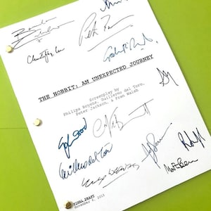 May include: A white script page from "The Hobbit: An Unexpected Journey" with multiple signatures in black ink. The title and screenplay credits are visible. The page is held together with gold brads against a green background.