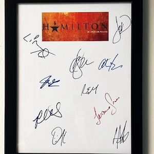 May include: A white framed print with black matting featuring the signatures of the cast of the musical Hamilton. The signatures are in black, blue, and red ink. The print also features a red, orange, and yellow logo for the musical Hamilton: An American Musical.