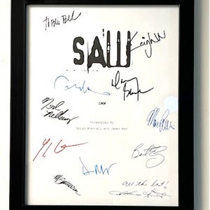 May include: A framed black and white script for the movie "SAW" with the title in bold and the names of the screenwriters Leigh Whannell and James Wan. The script is signed by several people, including Tobin Bell, Shawnee Smith, and Cary Elwes.