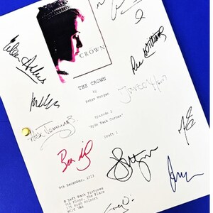 May include: A white script with the title "THE CROWN" and multiple signatures in black and red ink. The script features a profile image of a woman wearing a crown. The script is on a blue surface.