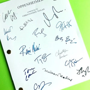 May include: A white script page for the movie "Oppenheimer" with the title and "screenplay by Christopher Nolan" printed on it. The page is signed by several people, including the director Christopher Nolan.