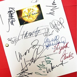 May include: A white script page from the movie "The Hunger Games" with multiple signatures in black, blue, green, and red ink. The signatures include "Jennifer Lawrence", "Elizabeth Banks", "Woody Harrelson", "Josh Hutcherson", and "Liam Hemsworth". The script page includes the title "The Hunger Games" and the production company "Colorforce/Monsgate Production" with the year "2012".