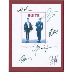 May include: A framed print featuring the word "SUITS" above an image of two men in suits. The print is surrounded by multiple signatures in blue ink and has a maroon border. The text "Created by Aaron Korsh" is at the bottom.
